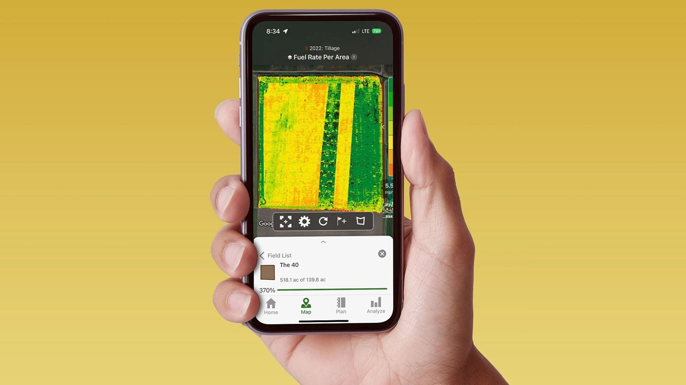 Egy kéz okostelefont tart, a képernyőn a John Deere Operations Center üzemanyag-felhasználással kapcsolatos adatai látszanak.