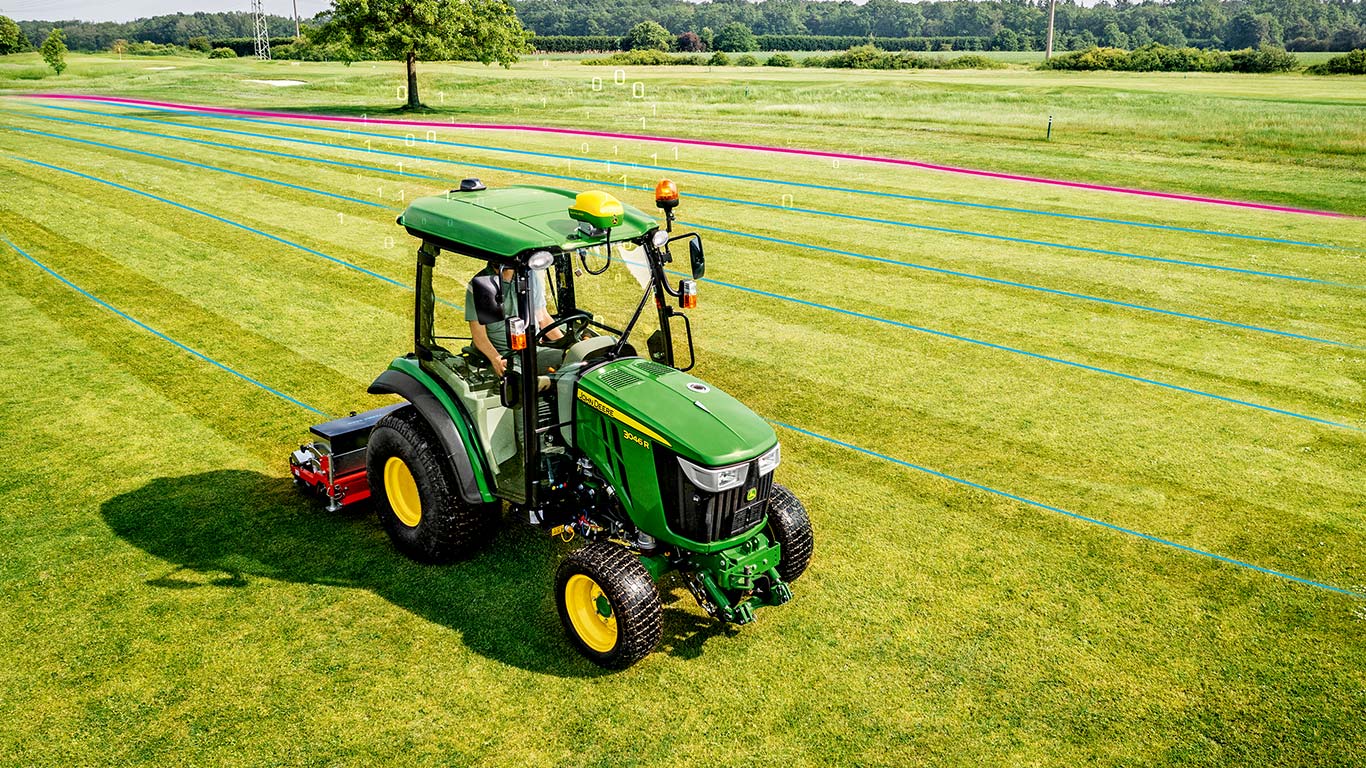 Egy golfpálya-karbantartó egy gyepterületen dolgozik egy John Deere kompakt többcélú traktorral.