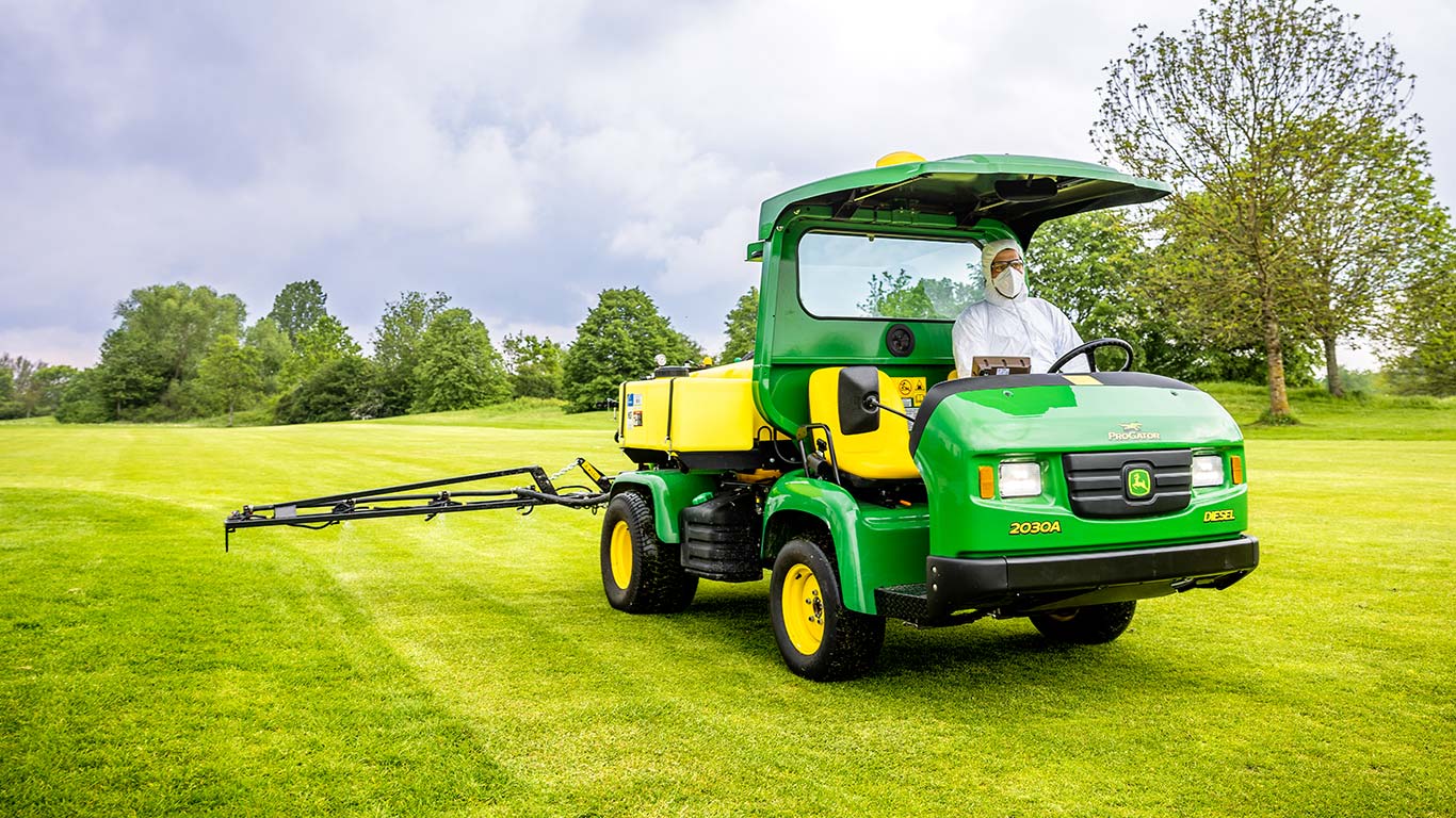 Egy John Deere-permetezőgéppel dolgozó, védőruhát viselő golfpálya-karbantartó egy golfpálya gyepén dolgozik.