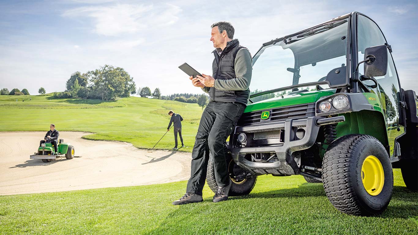 Egy vezető golfpálya-gondozó ellenőrzi az adatokat a táblagépén, miközben 2 golfpálya-gondozó dolgozik egy golfpályán a háttérben.
