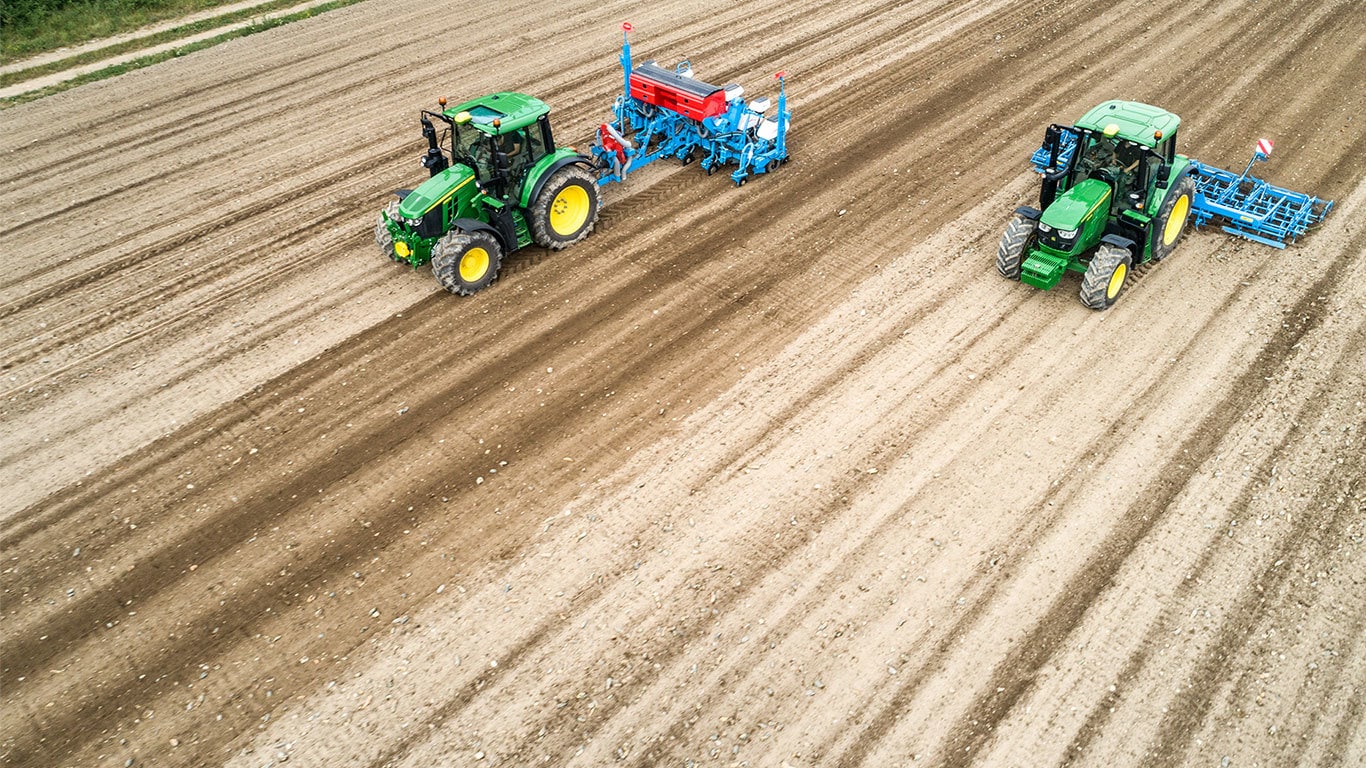 Két John Deere 6M traktor egy kopár földterületen. Az egyik egy Monsomen típusú szemenkénti vetőgépet húz maga után. A másik egy Lemken magágykészítő kombinációt vontat.