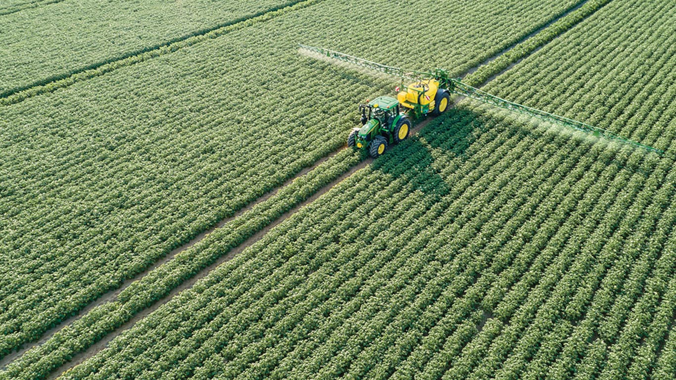 John Deere 6M sorozatú traktor M700 vontatott permetezőgépet húz maga után a burgonyaföldön.