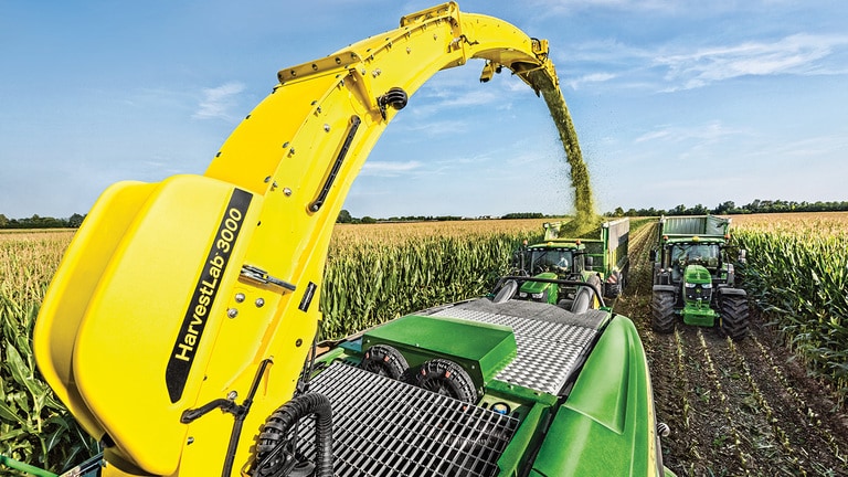 Kilátás egy John Deere 9000 sorozatú önjáró szecskázógép hátuljából, két silókocsival, amelyeket közvetlenül mögötte John Deere traktorok vontatnak.