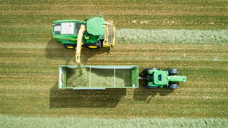 Légifotó a John Deere 8000 sorozatú, rendfelszedővel felszerelt önjáró szecskázógépről, amely a silótakarmányt John Deere traktor által vontatott kocsiba rakja.