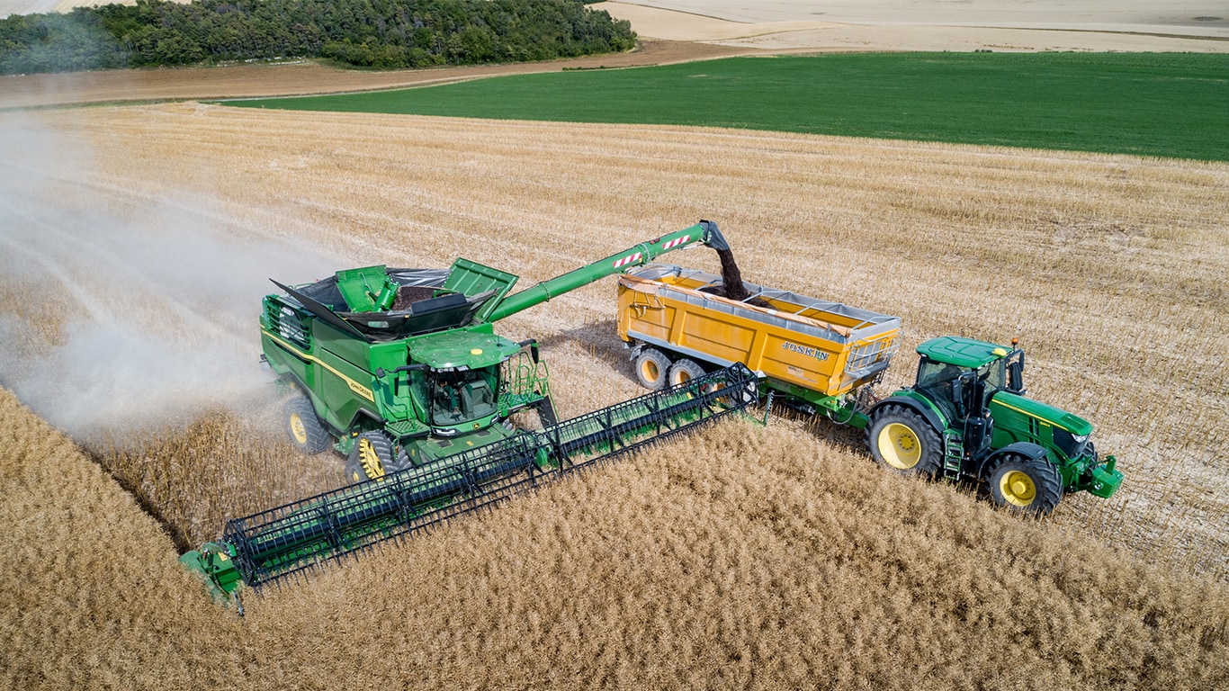 Egy John Deere X sorozatú kombájn repcemagot ürít egy John Deere traktorral vontatott magszállító kocsiba.