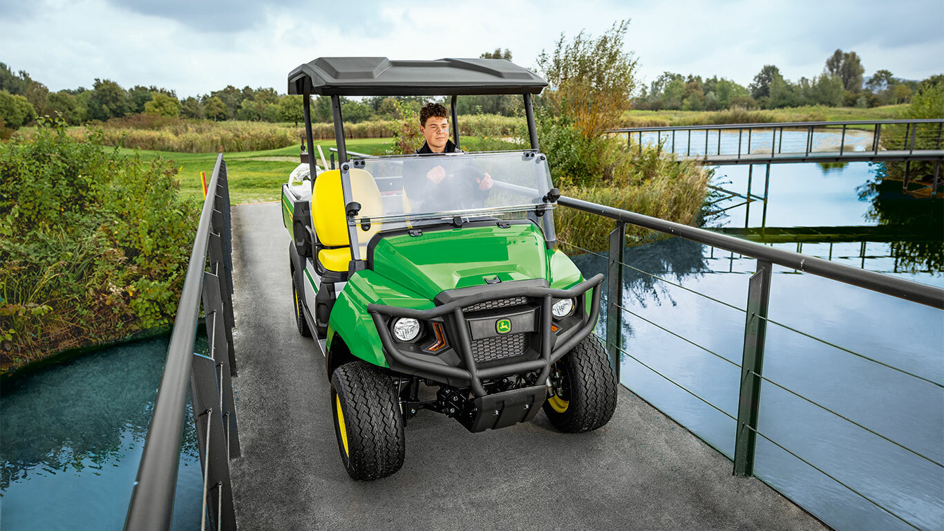 A John Deere Gator GS többcélú jármű egy keskeny hídon halad át egy nyugodt vízfolyás felett, amelyet buja zöld növények és fák vesznek körül. A John Deere Gator GS többcélú jármű egy keskeny hídon halad át egy nyugodt vízfolyás felett, amelyet buja zöld növények és fák vesznek körül.