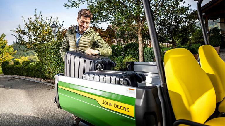 Egy nagy, fekete bőröndöt egy személy egy világos sárga ülésekkel szerelt John Deere Gator GS rakterébe helyez, zölddel és fákkal körülvéve.