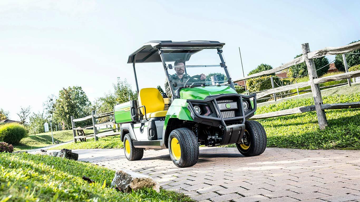 Zöld John Deere Gator GS többcélú jármű, amely burkolt útvonalon halad egy füves és fakerítéssel körülvett, parkosított területen.
