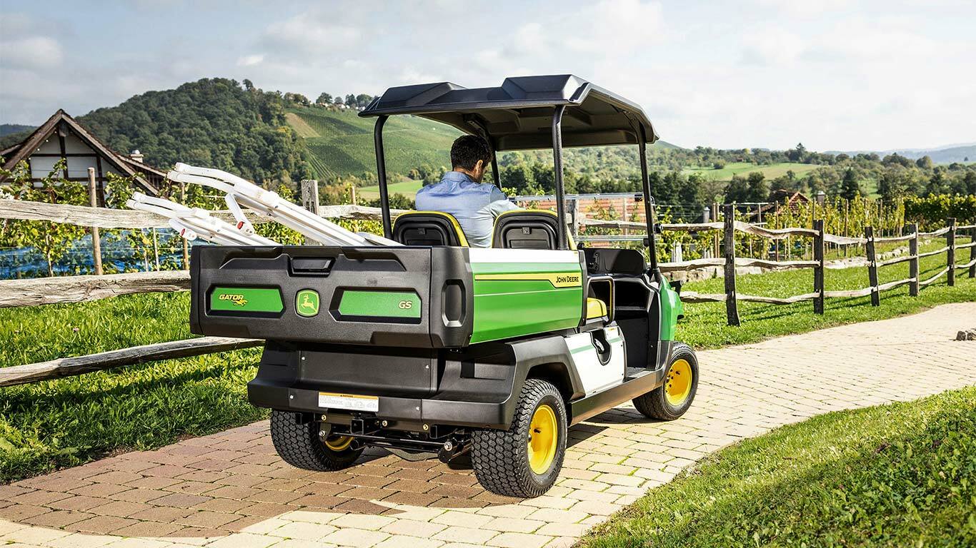 Hátulnézet egy John Deere Gator GS-ről, amely egy téglával burkolt úton halad, a háttérben festői dombokkal és szőlőültetvényekkel.