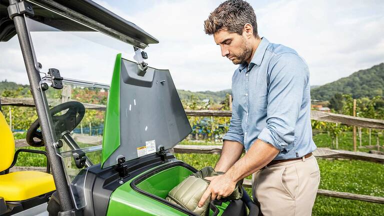 Egy párnázott zsákot egy zöld John Deere Gator GS elülső tárolórekeszébe helyez egy személy, amely a szabadban, egy fakerítés és egy szőlőültetvény közelében parkol.