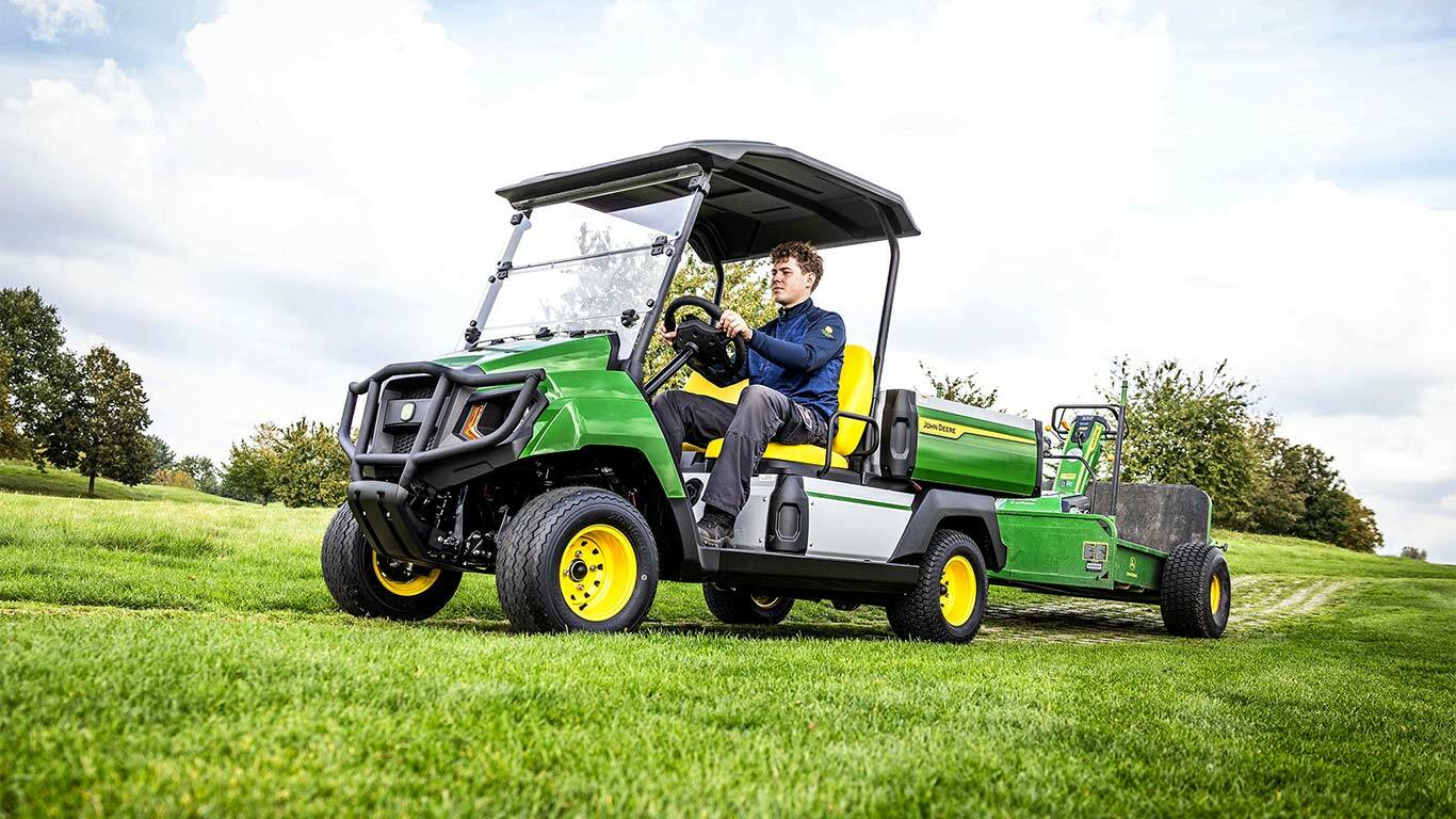 Zöld John Deere Gator elektromos többcélú jármű berendezést vontat egy füves területen egy részben felhős ég alatt.