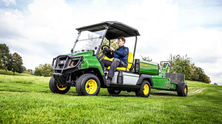 Zöld John Deere Gator elektromos többcélú jármű berendezést vontat egy füves területen egy részben felhős ég alatt.