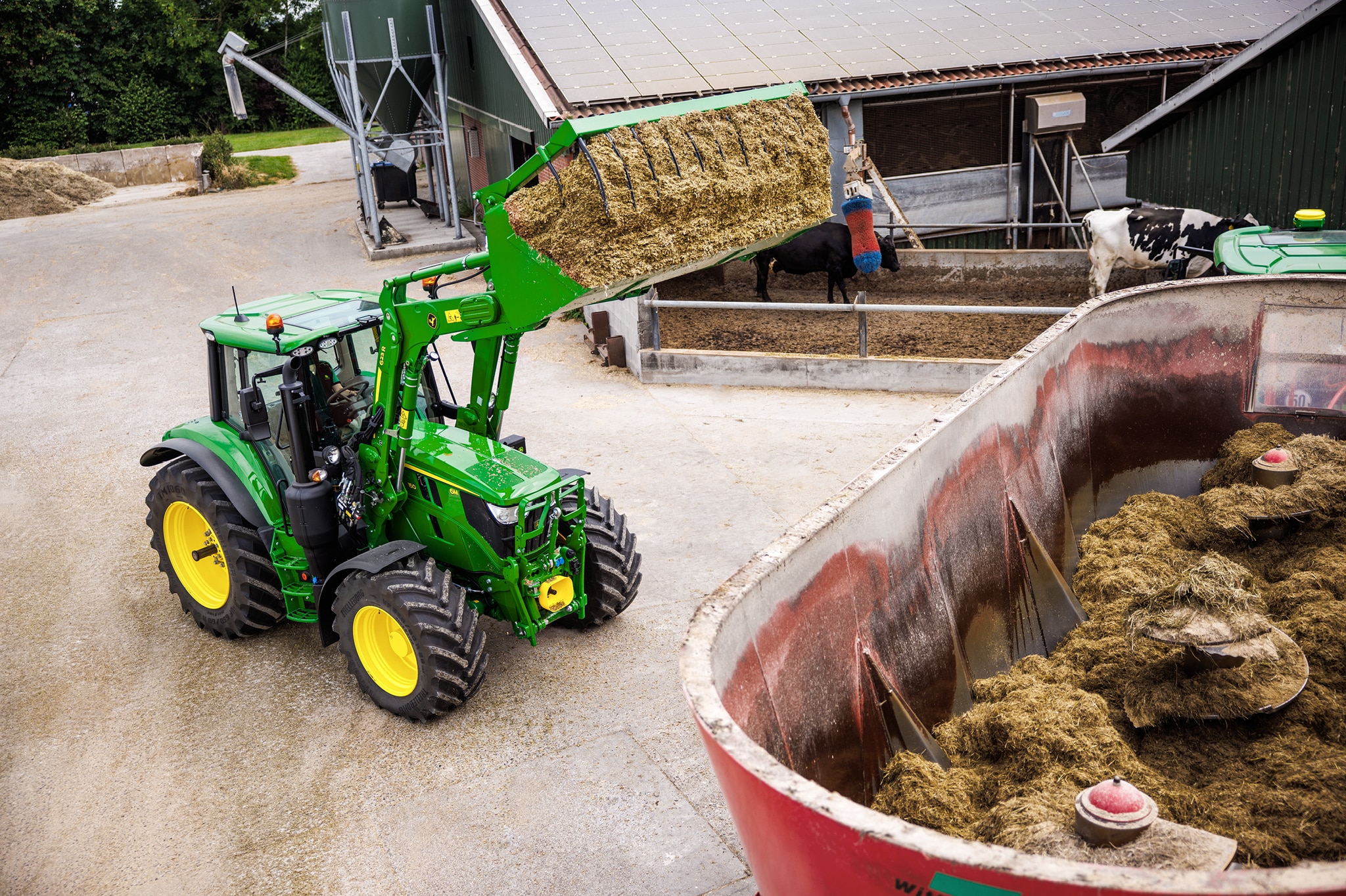 John Deere 6M sorozatú traktor homlokrakodóval silótakarmányt rakodik a gazdaság udvarán elhelyezett keverőkocsiba.