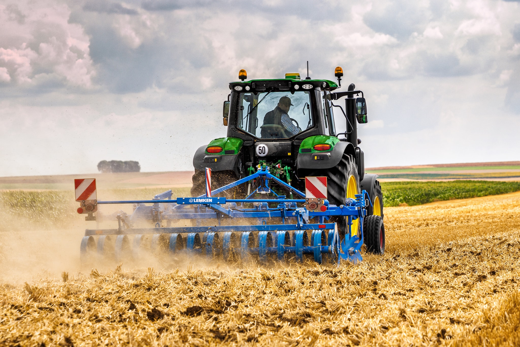 John Deere 6M sorozatú traktor, amely egy betakarított szántóföldön dolgozik tárcsás boronával, hátulnézetben poros szántóföldi körülmények között.