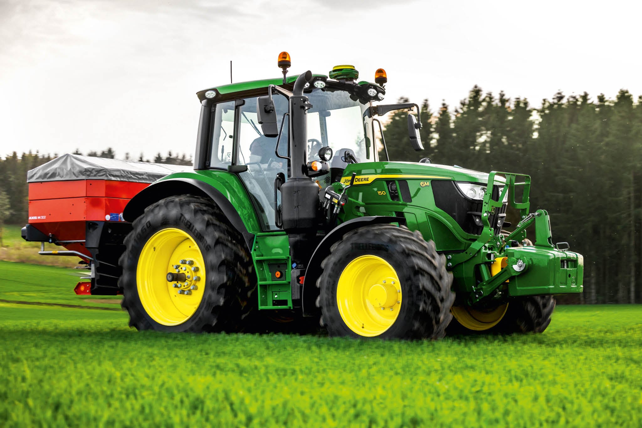 John Deere 6M sorozatú traktor hátsó függesztésű műtrágyaszóróval, amely az erdő szélén lévő fűben dolgozik.