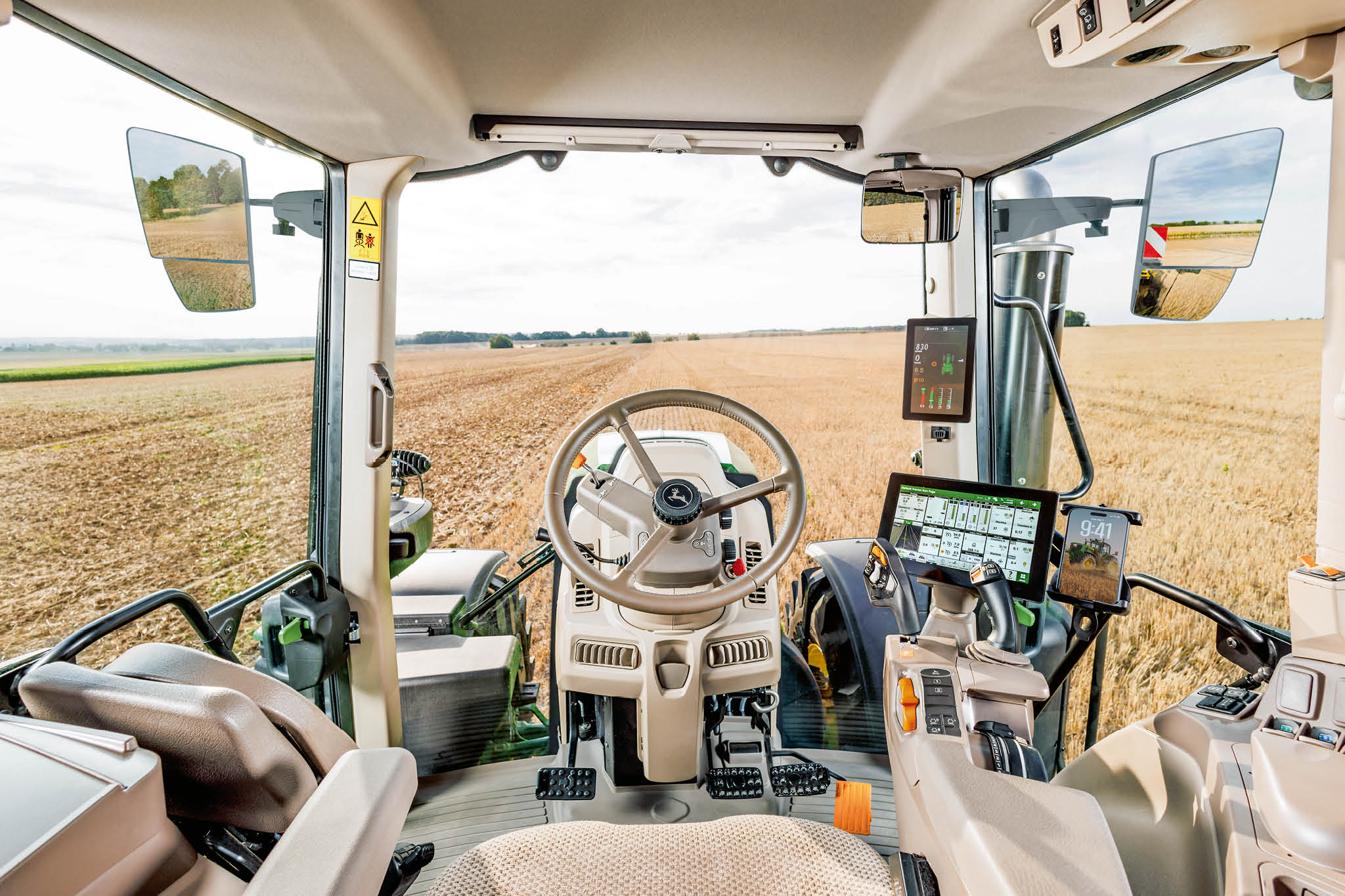 A John Deere traktor fülkéjének belseje kormánykerékkel, digitális kezelőszervekkel és a szélvédőn keresztüli szántóföldi rálátással a mezőgazdasági munka során.
