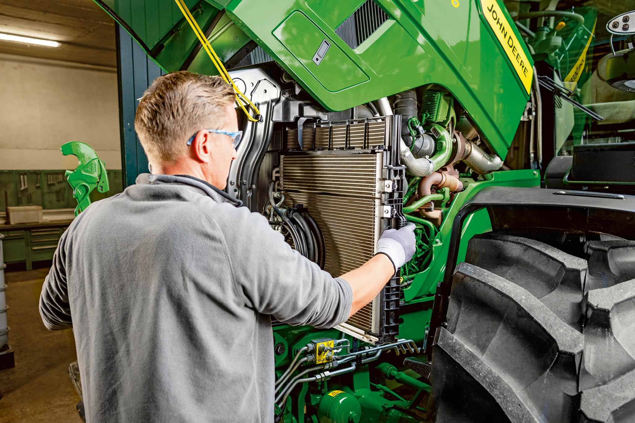 John Deere 6M sorozatú traktor motorterében karbantartást végző szerelő.