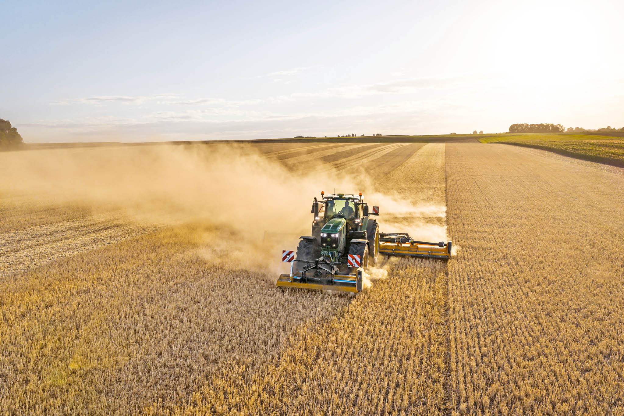 John Deere 6M sorozatú traktor hígtrágya tartálykocsit vontat fű és táblák által határolt országúton.