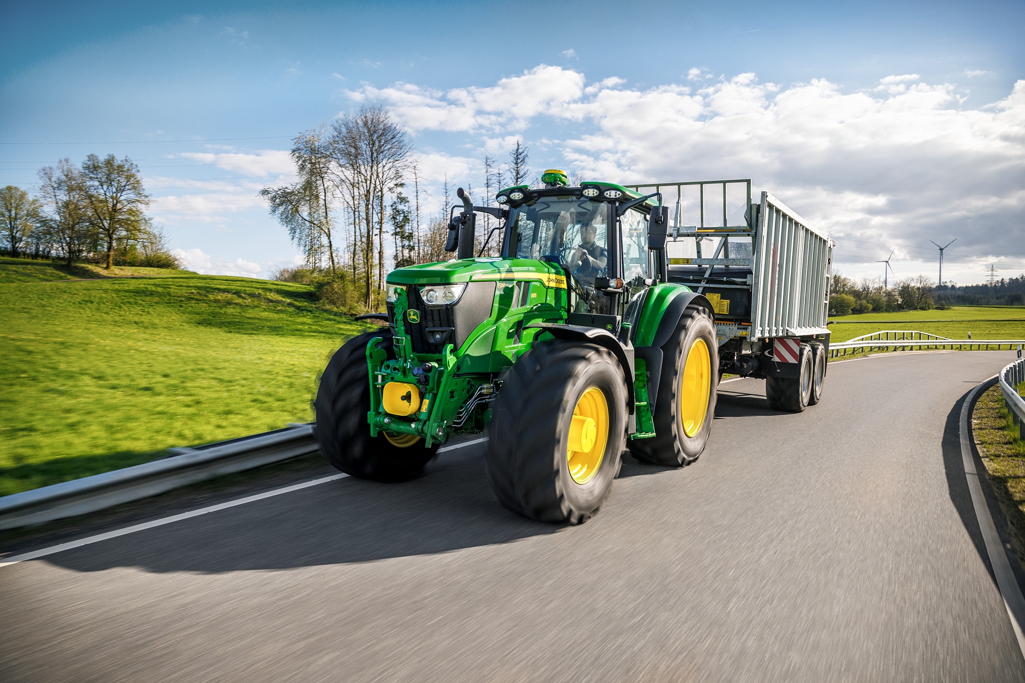 John Deere 6M sorozatú traktor magas oldalú pótkocsit szállít burkolt vidéki úton.