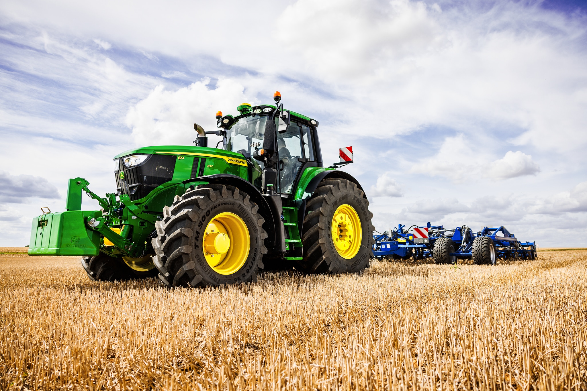 John Deere 6M sorozatú traktor nehéz talajművelő munkagépet vontat a tarlón, felhős égbolt alatt.
