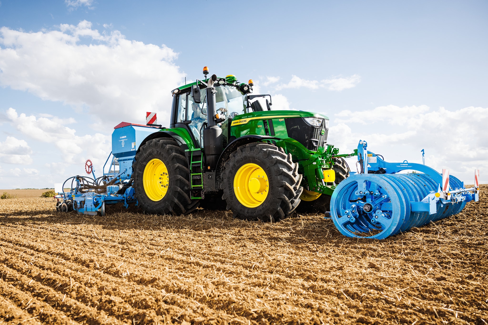 John Deere 6M sorozatú traktorok szántóföldön egy nagyméretű, kék munkagéppel.
