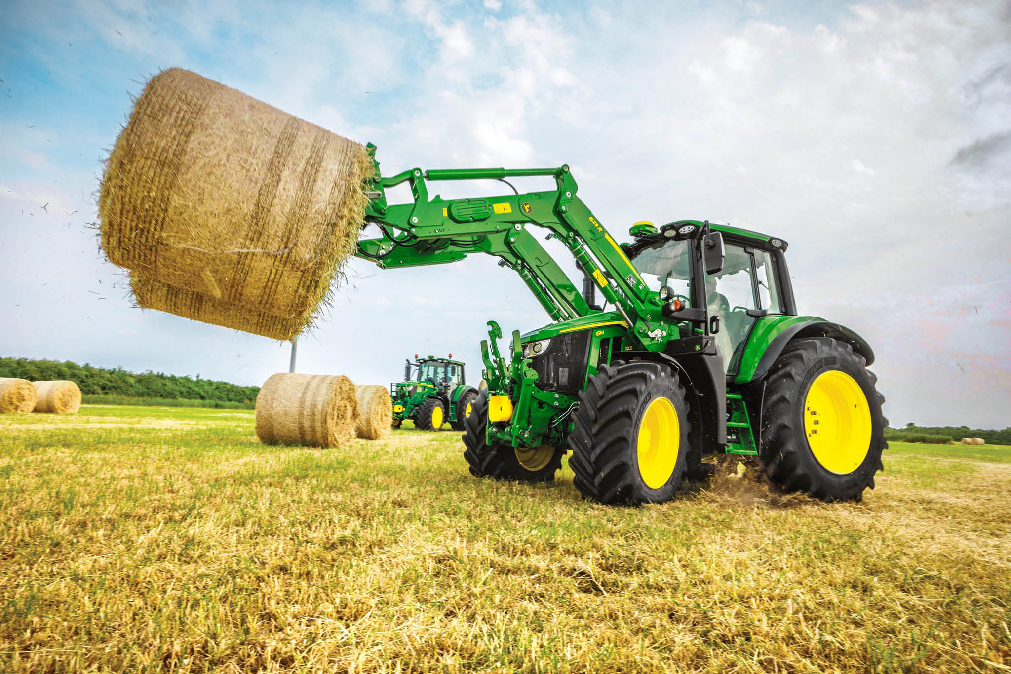 John Deere 6M sorozatú traktor egy nagy körbálát emel homlokrakodóval a nyílt terepen.