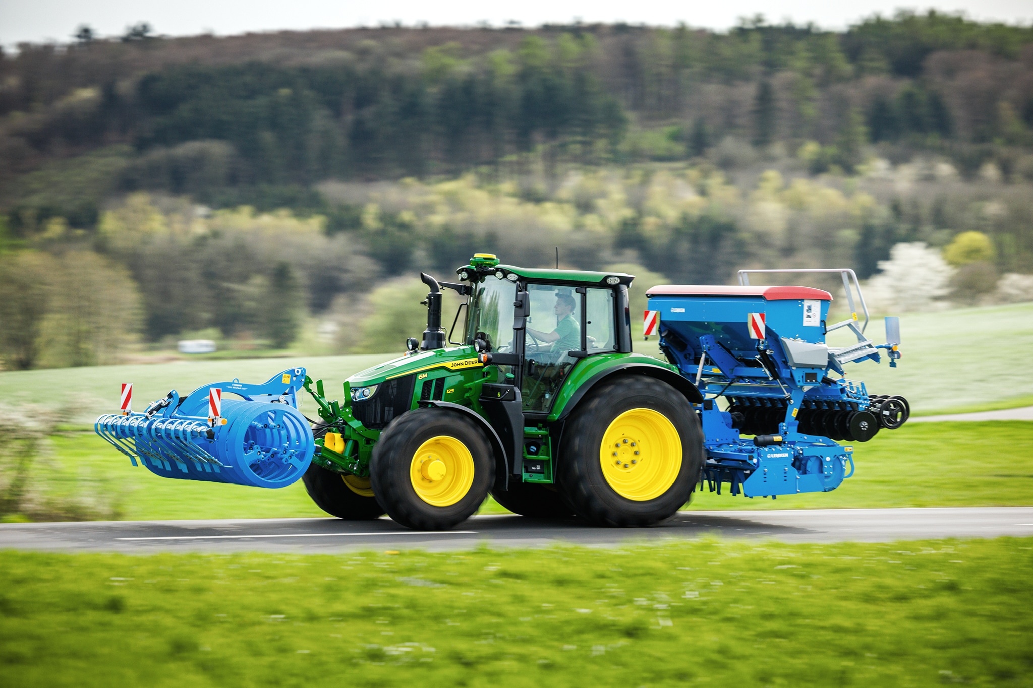 John Deere 6M sorozatú traktor egy mellső függesztésű munkaeszközt és hátsó vetőgépet szállít egy vidéki úton.