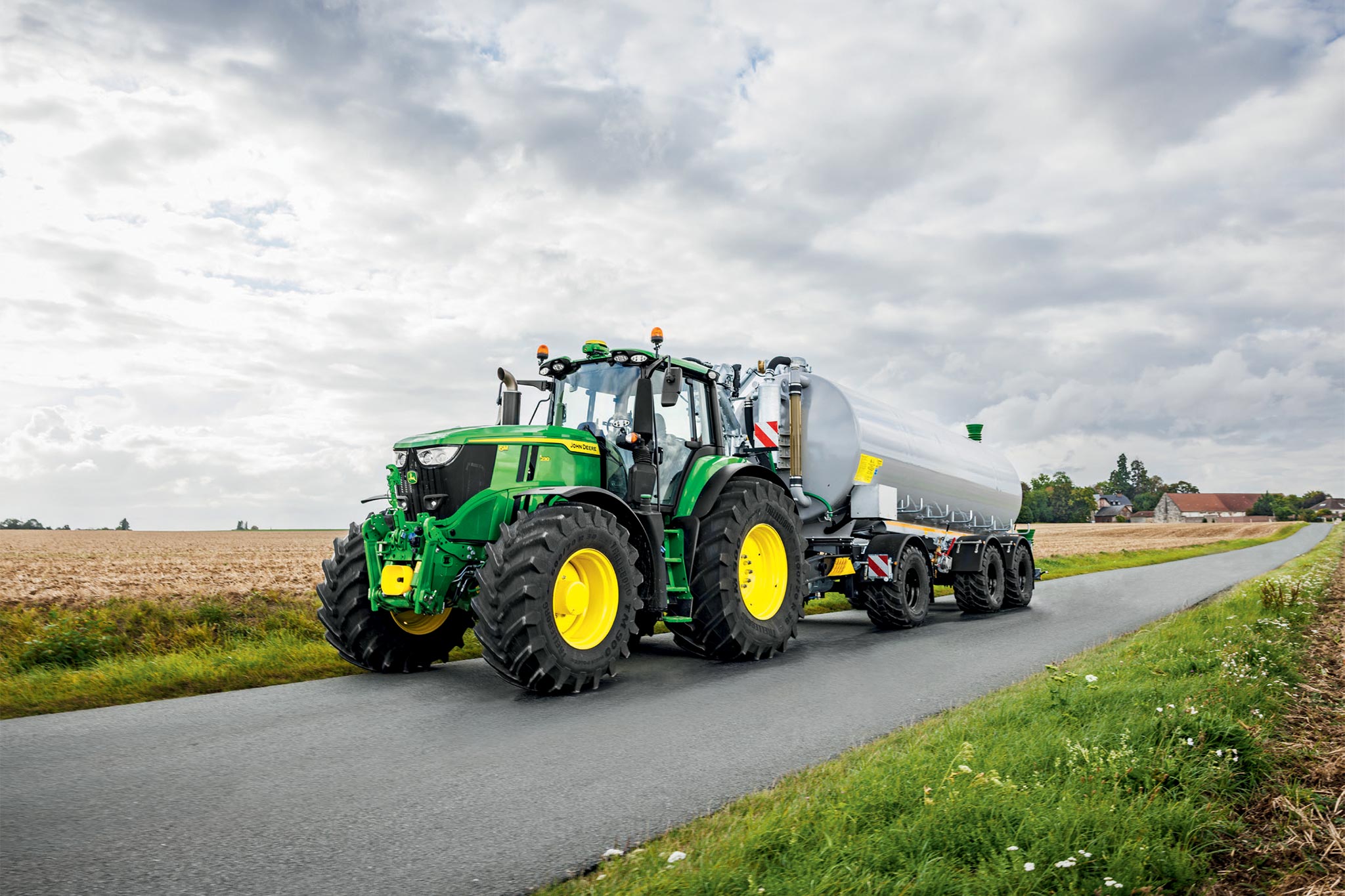 John Deere 6M sorozatú traktor széles kultivátorokkal dolgozik a szántóföldön, felülről nézve, szálló porral.