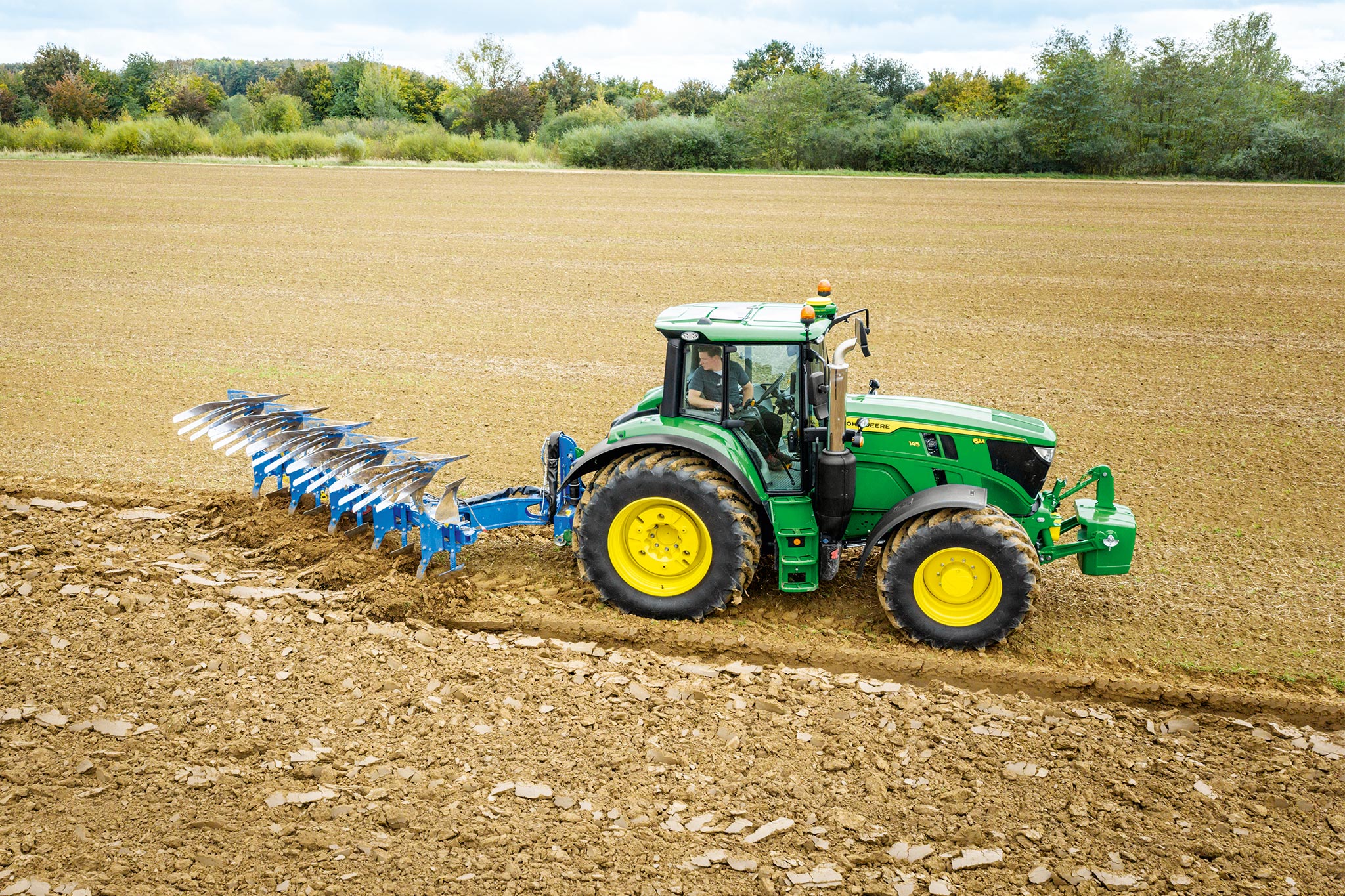 John Deere 6M sorozatú traktorszántott szántóföldön dolgozik hátsó talajművelő munkagéppel felhős égbolt alatt.
