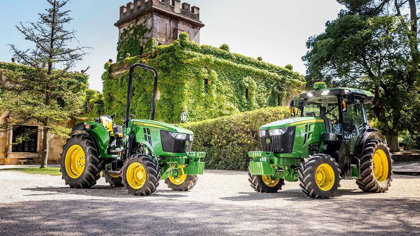Zöld és sárga John Deere 5075EN traktor, amely egy gyümölcsösön halad keresztül egy földúton, napsütéses gyümölcsfákkal körülvéve.