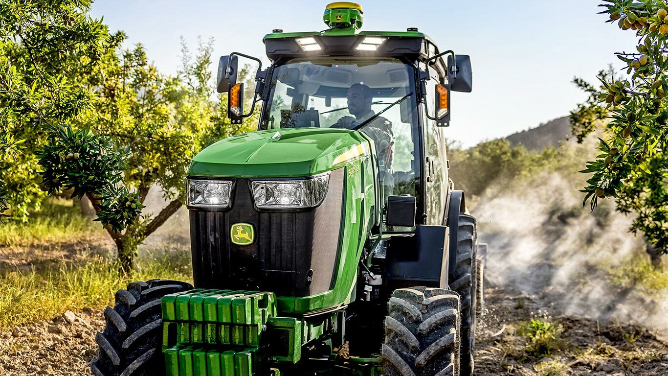 Zöld és sárga John Deere 5075EN traktor, amely egy gyümölcsösön halad keresztül egy földúton, napsütéses gyümölcsfákkal körülvéve.