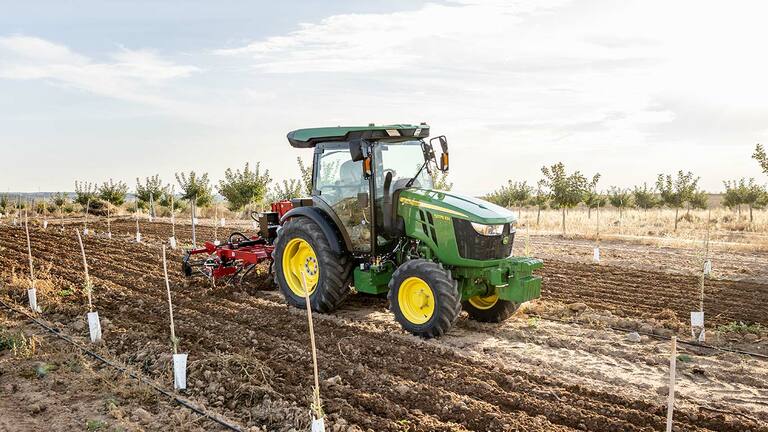 John Deere 5075EN traktor talajt művel nyílt mezőn tiszta ég alatt és ültetett sorok között.