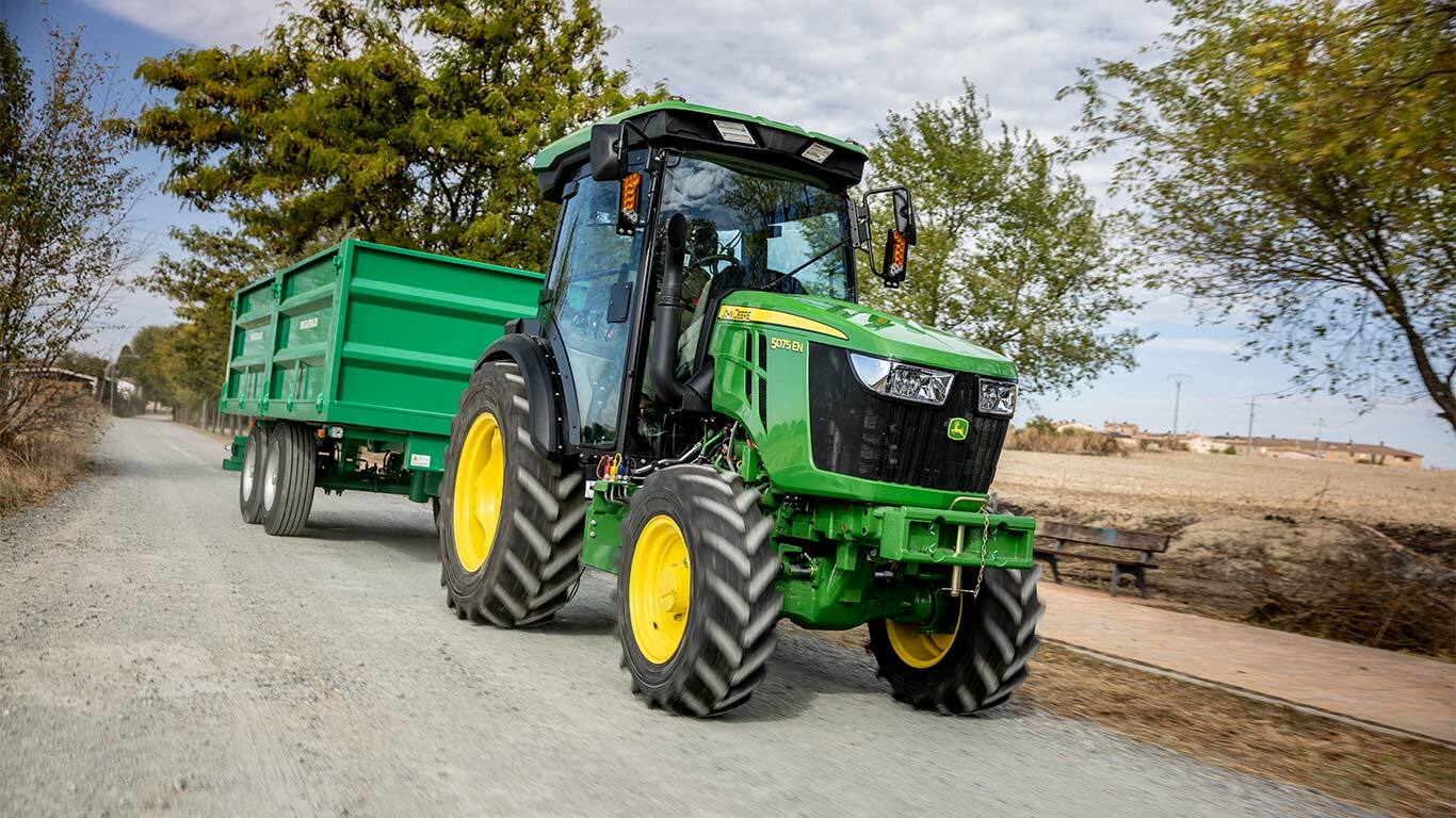 John Deere 5075EN traktor zöld pótkocsit húz vidéki földúton, háttérben fákkal.