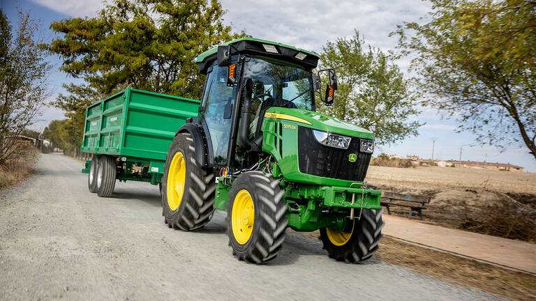 John Deere 5075EN traktor zöld pótkocsit húz vidéki földúton, háttérben fákkal.