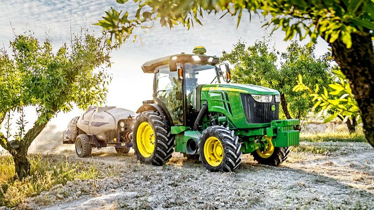 Zöld és sárga John Deere 5090EN traktor, amely egy permetezőgépet vontat egy gyümölcsösben egy napfényes napon.