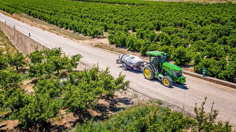 Zöld és sárga John Deere 5090EN traktor, amely permetezőgépet vontat egy burkolt úton a nagy gyümölcsös sorok között, a tiszta kék ég alatt.