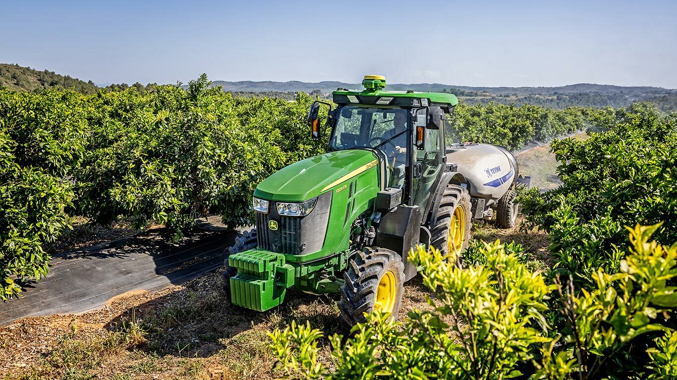 Zöld és sárga John Deere 5105EN traktor, amely egy permetezőgépet vontat egy jól fejlett gyümölcsösben, a háttérben hegyekkel.