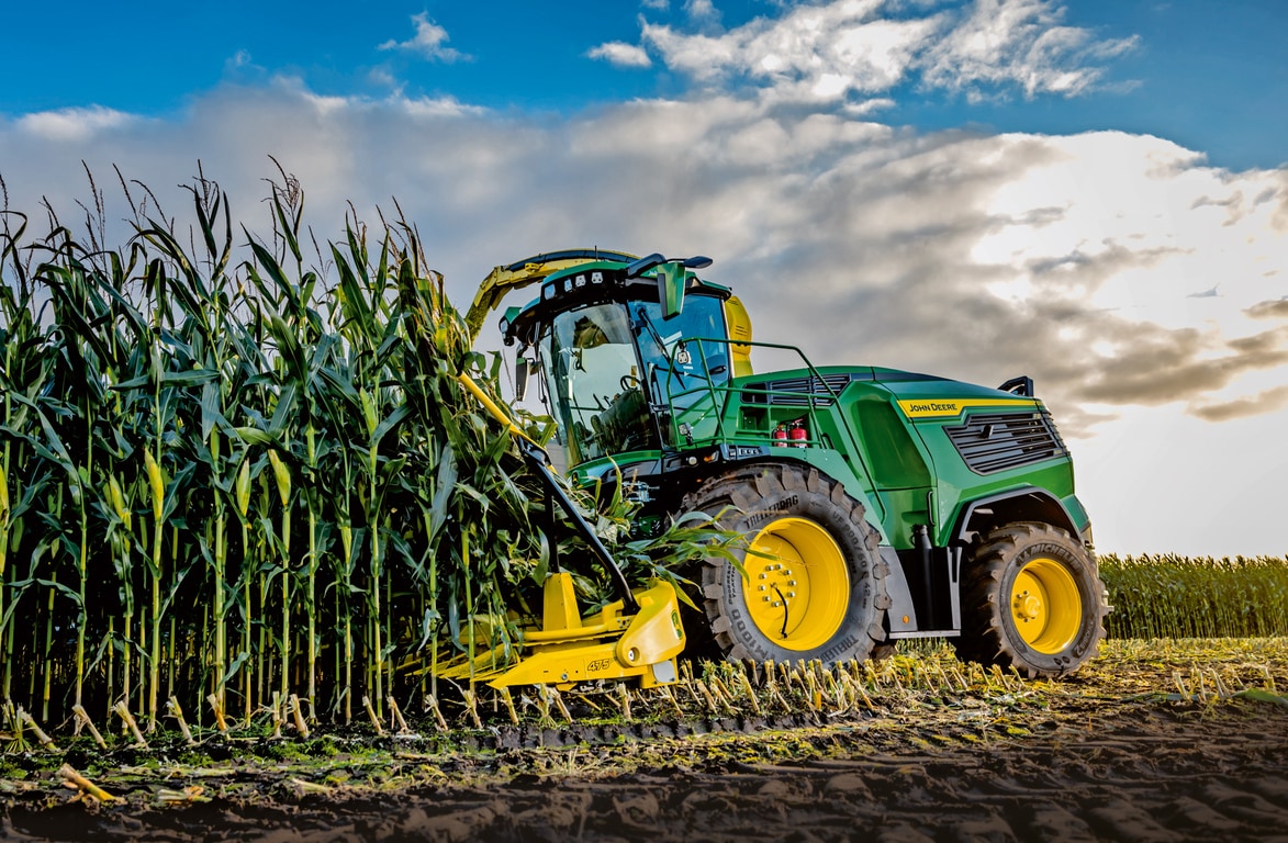 John Deere önjáró szecskázó kukoricát takarít be egy szántóföldön, kék felhős égbolt alatt.