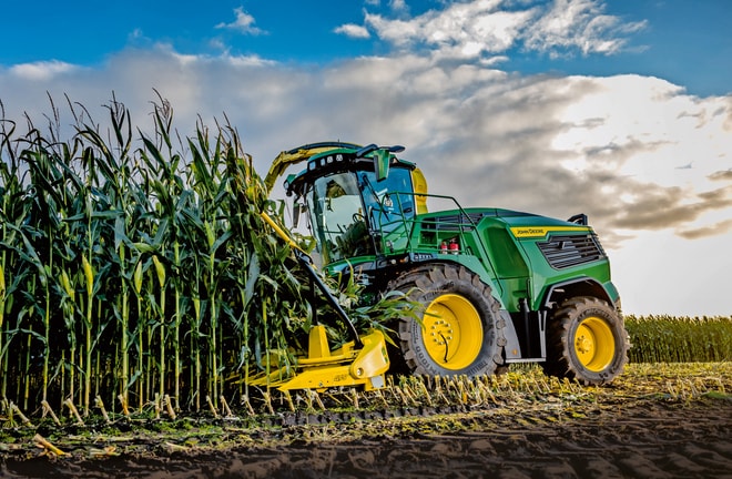 John Deere önjáró szecskázó kukoricát takarít be egy szántóföldön, kék felhős égbolt alatt.