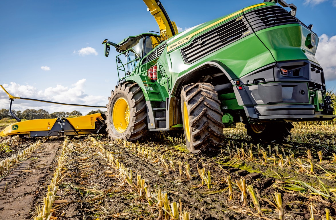 John Deere önjáró szecskázó dolgozik egy betakarított kukoricaföldön, bemutatva a gép nagyméretű gumiabroncsait és vágószerkezetét a tiszta kék égbolt előtt.