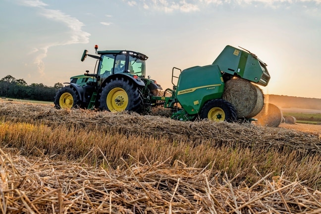 Egy John Deere 6M traktor által vontatott John Deere F442M bálázó szalmabálát dob ki