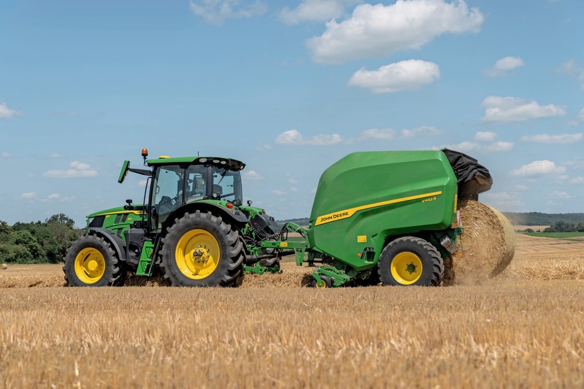 Egy John Deere 6R 150 által vontatott John Deere V452R egy szalmás tarlón bálát dob ki