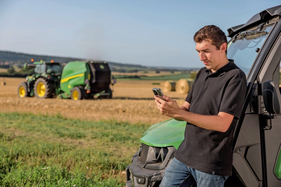 A bála dokumentációs adatait a mobiltelefonon ellenőrző személy egy John Deere VR bálázóval a háttérben