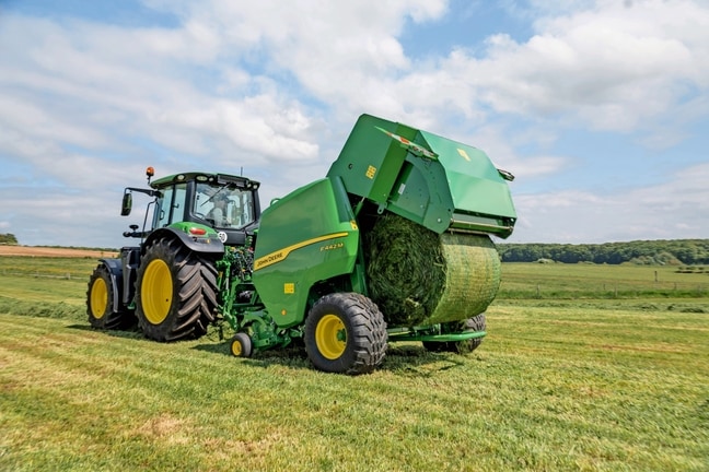 Egy John Deere 6M traktor által vontatott John Deere F442M bálázó fűszéna bálát dob ki