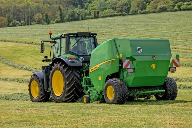 Egy John Deere 6M traktor által vontatott John Deere F442M bálázó egy dombos füves területen