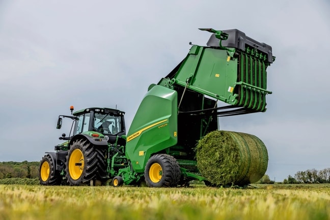 Egy John Deere 6M traktor által vontatott John Deere V462M bálázó fűszéna bálát dob ki